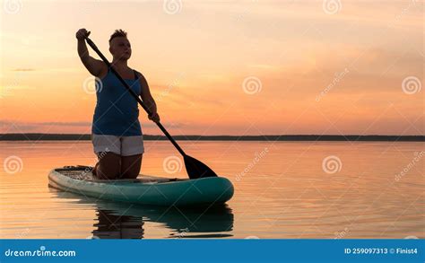 A Woman With A Mohawk In Shorts On Her Knees On A Sup Board With An Oar