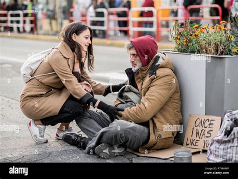 Young Woman Giving Money To Homeless Beggar Man Sitting In City Stock