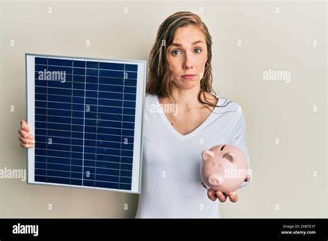 Jeune femme blonde tenant panneau solaire photovoltaïque et portique banque sceptique et nerveux