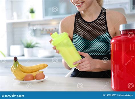 Young Woman Holding Bottle Of Protein Shake At Table With Ingredients