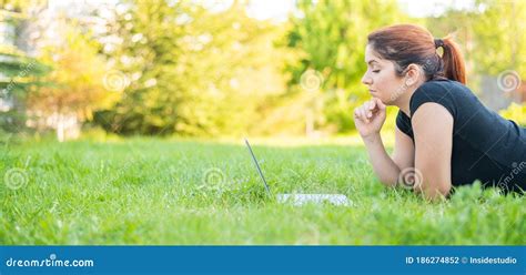 A Woman Lies On Her Stomach In A Park And Types On A Laptop Female