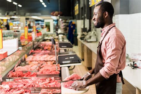 Side View Of Smilng African American Male Butcher In Apron Cutting Raw