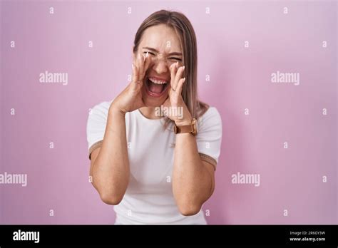 Blonde Caucasian Woman Standing Over Pink Background Shouting Angry Out Loud With Hands Over