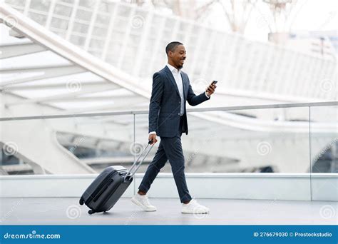 Handsome Black Businessman Walking With Suitcase At Airport And Using