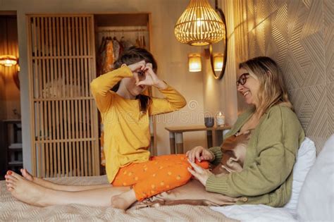 An Attractive Blonde Mom With Glasses And Her Cute Daughter Are Sitting On The Bed And Smiling