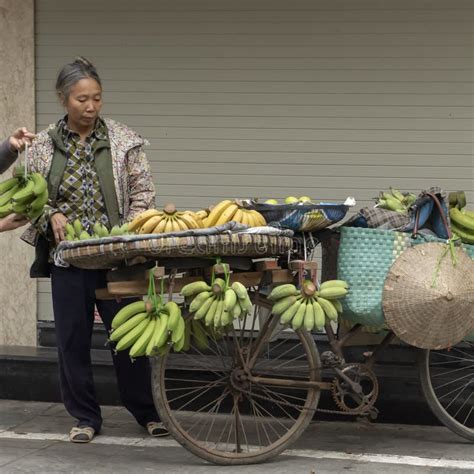 Vietnamese Street Vendor In Hanoi Walking Across A Street Carrying Flowers To Sell In Each Hand