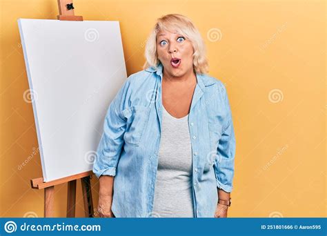 Middle Age Blonde Woman Standing By Painter Easel Stand Afraid And