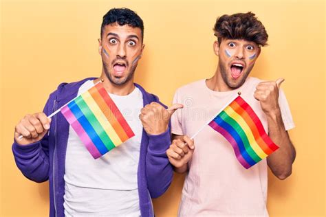 Young Gay Couple Holding Rainbow Lgbtq Flags Pointing Thumb Up To The