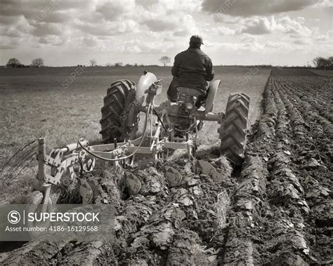 1950s BACK VIEW OF ANONYMOUS FARMER ON TRACTOR PLOWING A FIELD TURNING ...