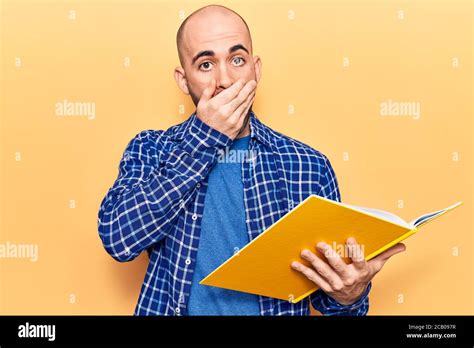 Young Handsome Bald Man Reading Book Covering Mouth With Hand Shocked