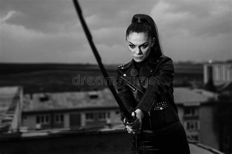 Grayscale Of Female In Black Ninja Assassin Costume Holding A Sword And Standing Near Wall Stock