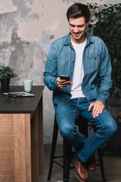 Hombre joven sonriente que se sienta en taburete usando el teléfono