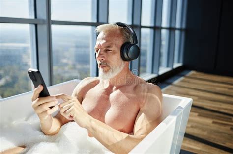 Mature Man Using Phone Listening To Music In Headphones While Lying In Bathtub Indoors Stock