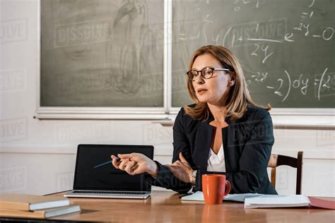 Female Teacher Sitting At Desk And Pointing At Laptop With Blank Screen