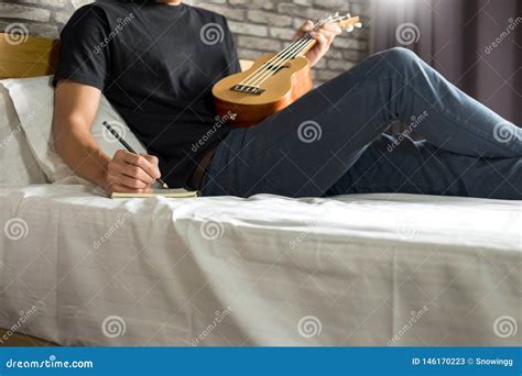 Happy Young Asian Man Playing Ukulele Sitting On Bed In Bedroom Stock