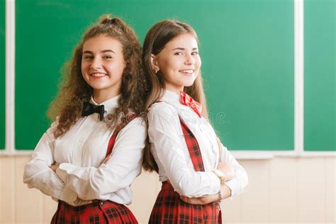 Two Brunette Schoolgirls In School Red Uniforms Are Standing In A Classroom With Books Stock
