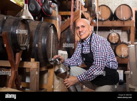 Smiling Mature Seller Pouring Wine From Wood Barrel In Wine Cellar