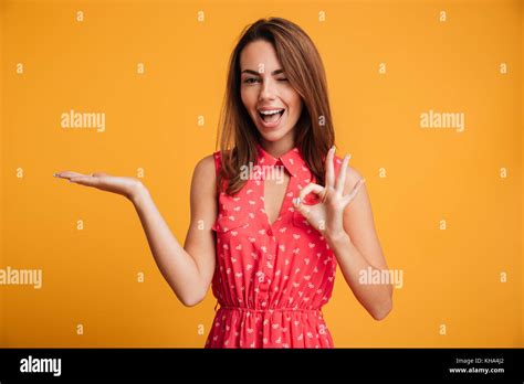 Happy Brunette Woman In Dress Holding Copyspace On The Pound And Showing Ok Sign While Looking