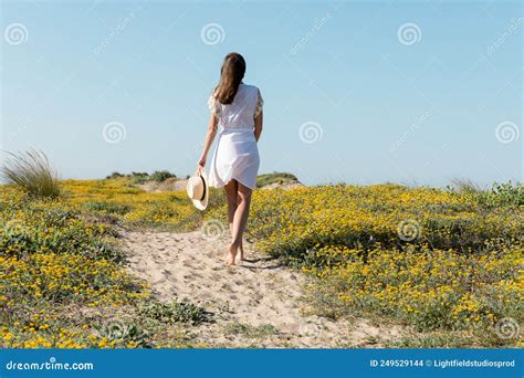 Back View Of Barefoot Woman Holding Straw Hat While Walking On Beach