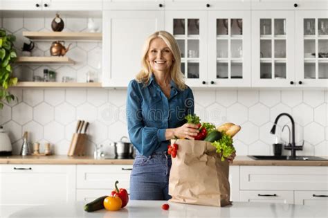 Happy Mature Woman Unpacking Paper Bag With Fresh Organic Vegetables In Kitchen Stock Photo