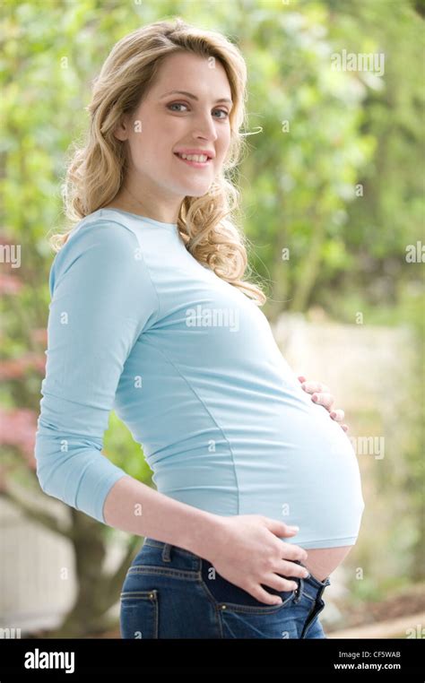 Pregnant Female Wavy Blonde Hair Wearing A Blue Top Long Sleeves And Jeans Standing In The