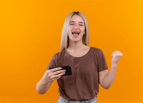 Joyeuse Jeune Fille Blonde En Appareil Dentaire Tenant Un Téléphone Mobile Avec Le Poing Levé Et