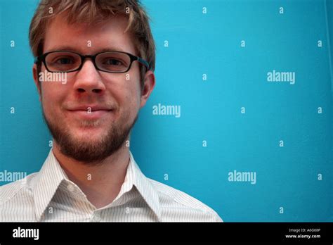 Young Bearded Man With Glasses Stands Smiling By A Blue Wall Stock