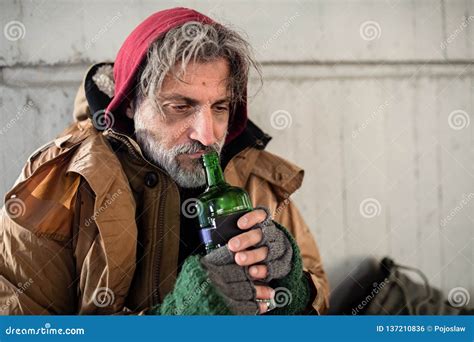 A Front View Of Homeless Beggar Man Sitting Outdoors Holding Bottle Of