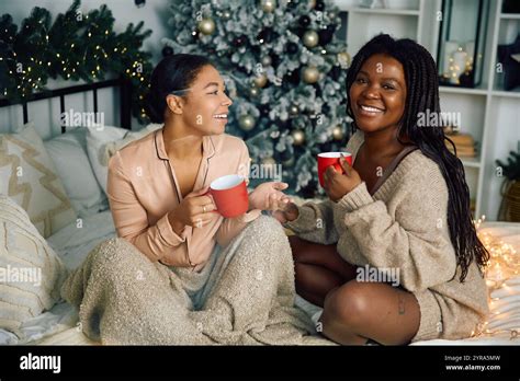 A Happy Lesbian Couple Enjoys Warm Drinks Together In Their Festive Cozy Bedroom Stock Photo