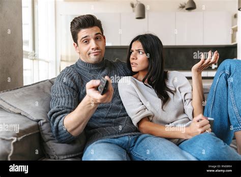 Bored Young Couple Sitting On A Couch At Home Watching Tv Drinking