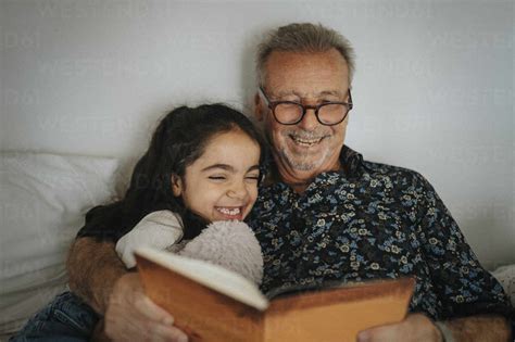 Smiling Senior Man Reading Storybook With Granddaughter On Bed At Home