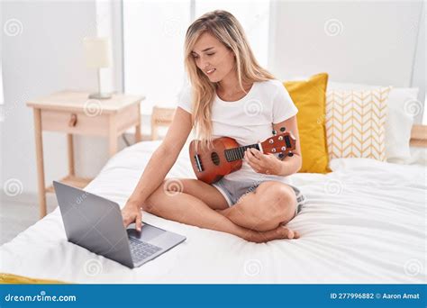 Young Blonde Woman Having Online Ukulele Lesson Sitting On Bed At