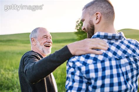 An Adult Hipster Son With Senior Father On A Walk In Nature At Sunset