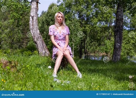 Young Beautiful Blonde Girl In A Pink Dress Sits On A Stump In A Forest