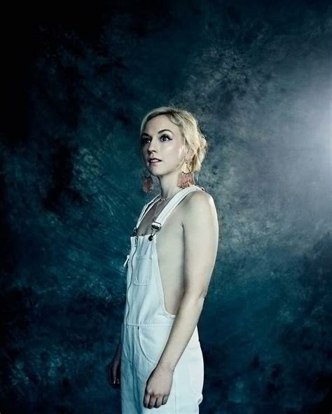 A Woman Standing In Front Of A Dark Background Wearing White Overalls