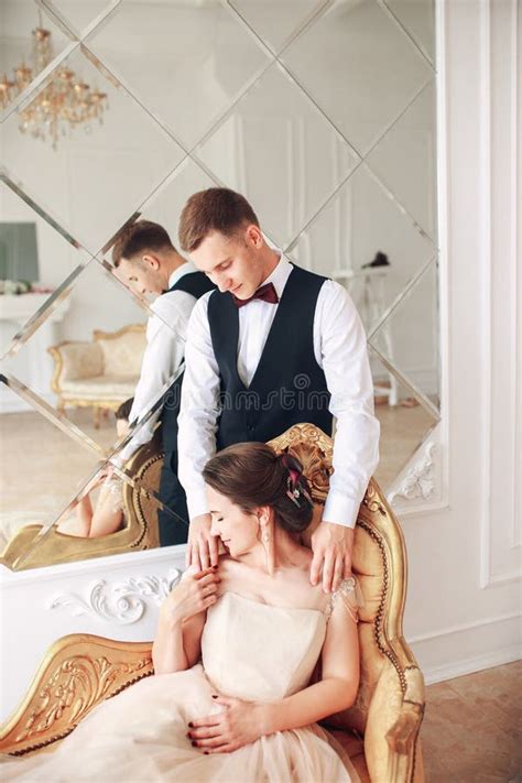 Bride In Beautiful Dress And Groom In Black Suit Sitting On Sofa