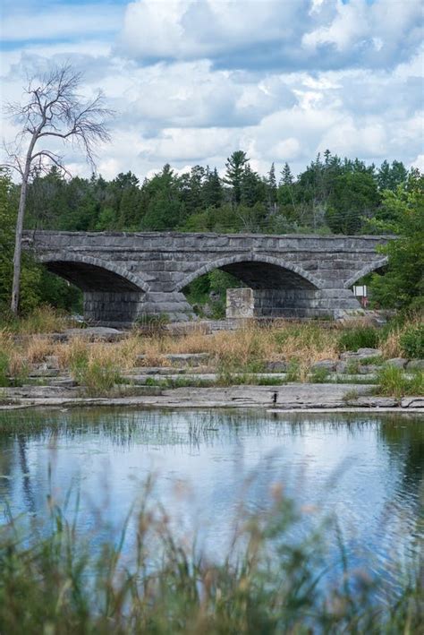 Arched Stone Made Bridge Over A Flowing Stream Of Water Surrounded By