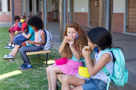 Two Girls Eating Lunch From Tiffin Box While Sitting On Bench In The Park At School Stock Image