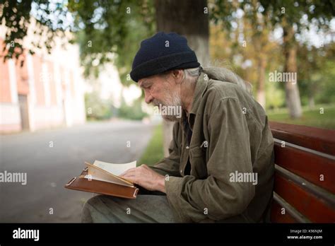 Side View Of Old Homeless Man With Long Grey Hair Reading A Book Stock