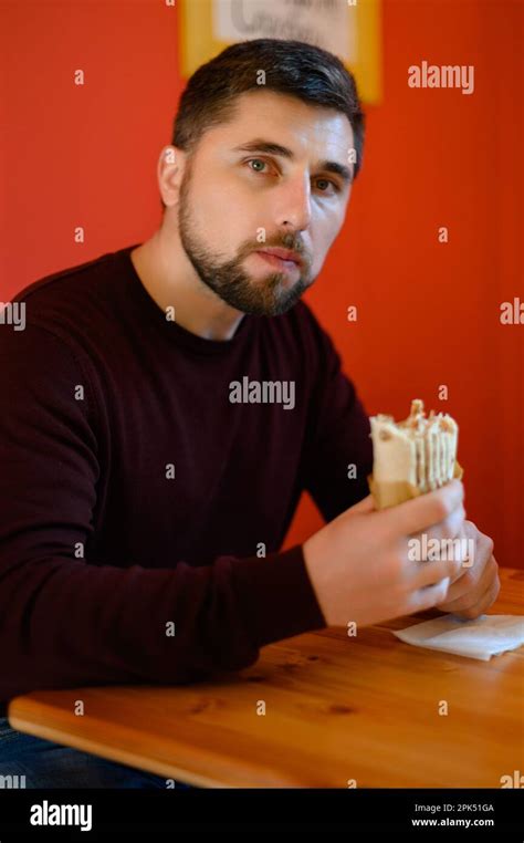 A Man With A Beard Is Sitting On A Chair And Eating Shawarma A Quick