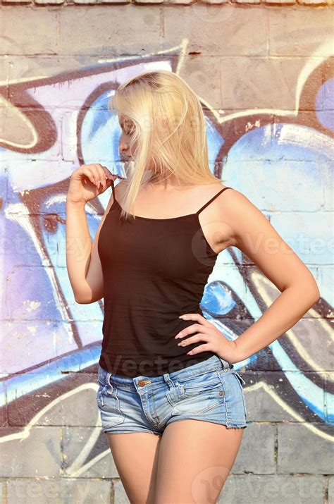 Sexy Caucasian Blonde Girl In Denim Shorts And Black Tank Top Posing Against Graffiti Wall In