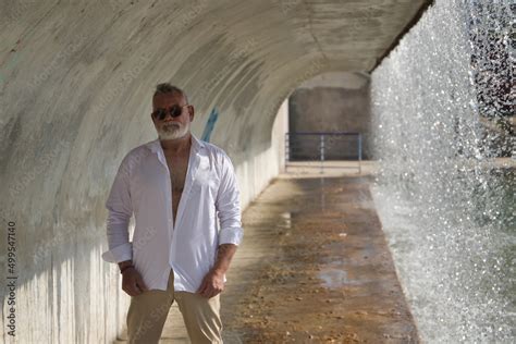 Mature Gray Haired Man With Beard Sunglasses Open White Shirt And Light Brown Pants Under A