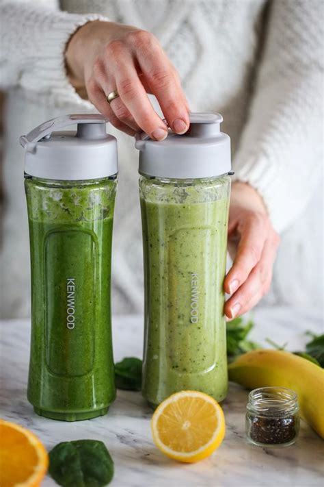 Two Blenders Filled With Green Smoothies Next To Bananas And Oranges