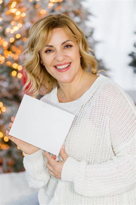 Blonde Woman Sitting In Home Christmas Setting And Holding White Sheet Of Paper Free For Text