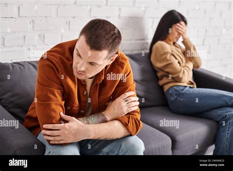 Upset Tattooed Man Sitting On Couch In Living Room Near Crying Asian