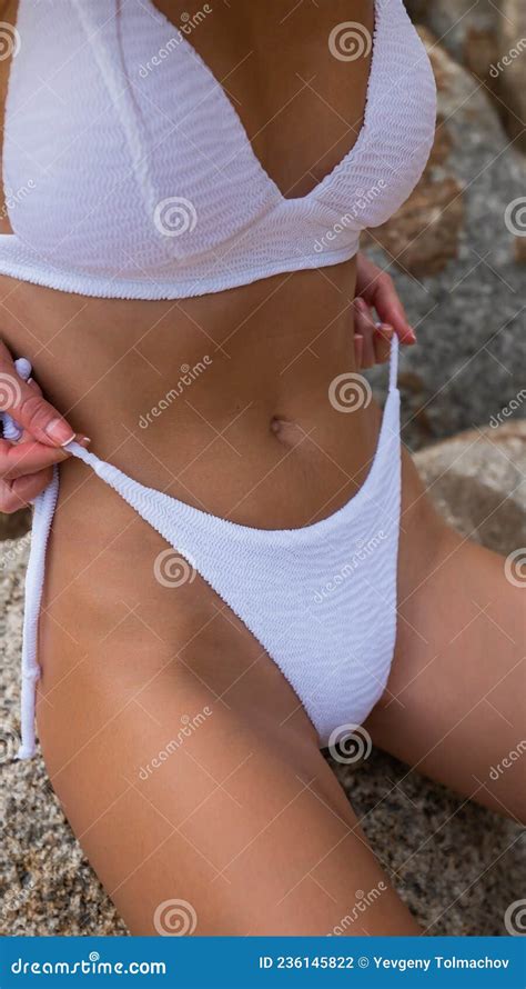Closeup Shot Of Slender Tanned Model In White Bikini Sitting On Stone Holding Her Thong With