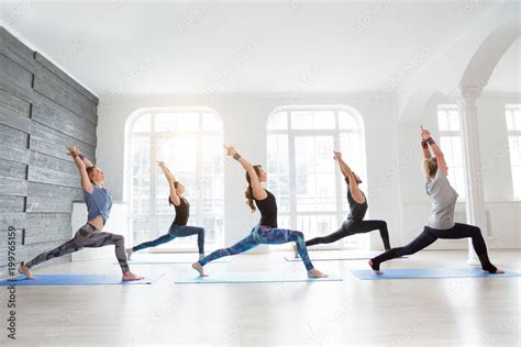 Group of people doing yoga warrior pose at white studio with gesture of will. Fitness class ...