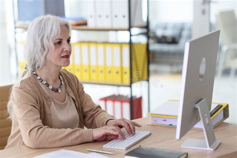 Positive Mature Employee Works On Computer At Desk In Office Stock