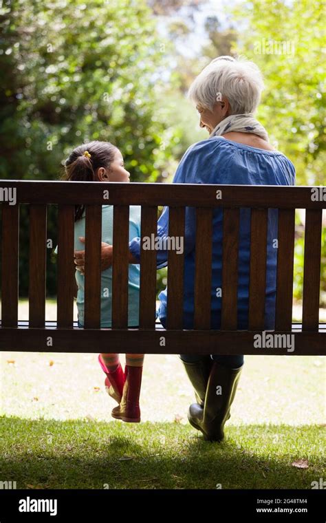 Rear View Of Grandmother And Granddaughter Sitting On Wooden Bench