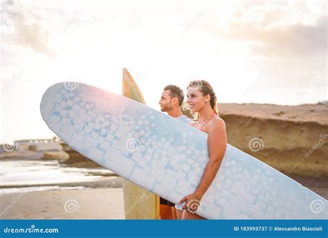 Young Couple Of Happy Surfers Standing On The Beach Holding Surfboards